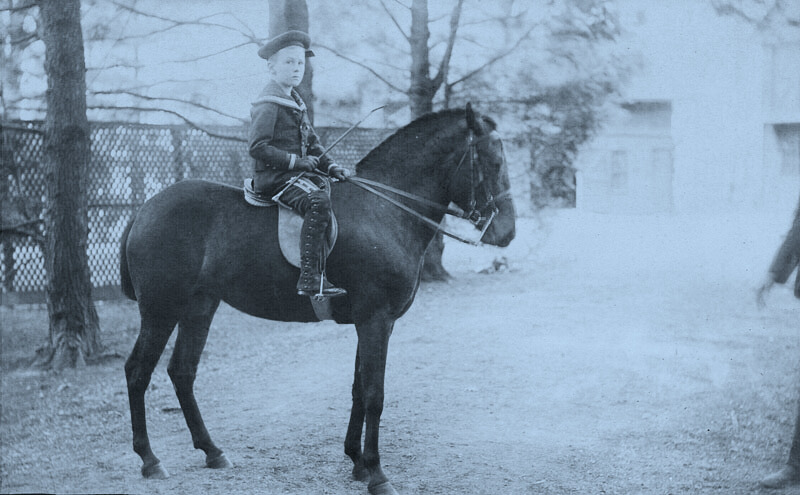 black-and-white photo of 10-year-old boy on a horse. He is holding a crop and looking at the camera; time period is about early 20th century. HOrse is standing on an unpaved road with a fence and stone building in the background.